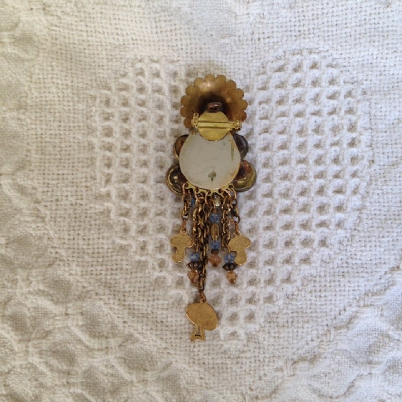 May include: A gold-toned brooch with a large, round, iridescent center surrounded by smaller, colorful stones. The brooch has a dangling chain with small, gold-toned charms.