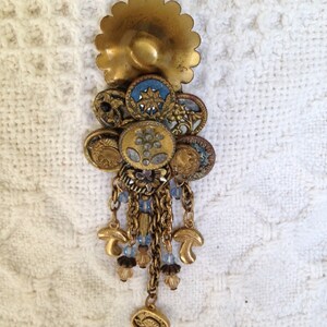 May include: A gold-toned pendant with a large, round, decorative top and a long, dangling chain of smaller buttons, beads, and charms. The pendant features a variety of textures and patterns, including floral motifs and geometric shapes.