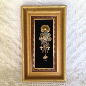 May include: A gold framed picture with a black background featuring a gold and blue jeweled pendant with a flower design.