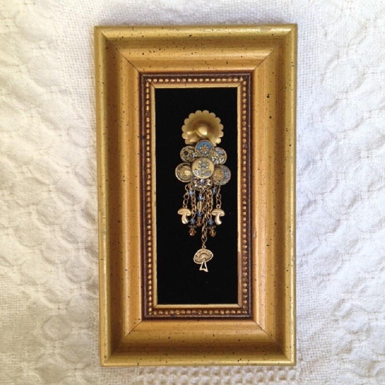 May include: A gold framed picture with a black background featuring a gold and blue jeweled pendant with a flower design.