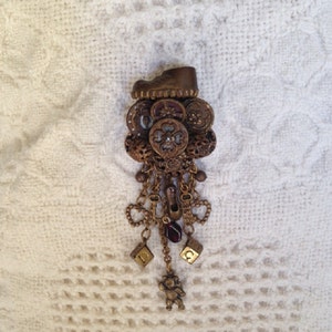 May include: A gold-toned brooch with a shoe charm, multiple buttons, and a dangling charm with a teddy bear. The brooch has a red gem and letter charms.