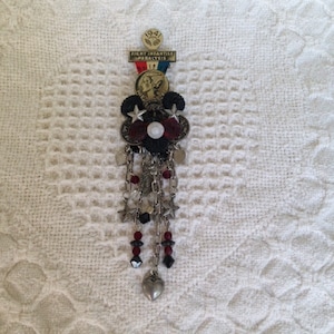 May include: A vintage brooch with a patriotic theme. The brooch features a banner that reads "FIGHT INFANTILE PARALYSIS" and the year "1941". It has a central design with stars, beads, and dangling chains, with a heart-shaped charm at the bottom.