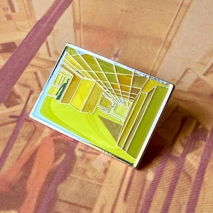 May include: A rectangular enamel pin featuring a stylized perspective illustration of an interior space with yellow, lime green, and white geometric lines. The design depicts a series of angular planes and grid-like structures in shades of yellow.
