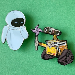 May include: Two enamel pins featuring characters from the animated film WALL-E. The first pin is a white robot with a blue face and a purple flower. The second pin is a yellow robot with a black and brown tread design and the text "WALL-E" on the side.