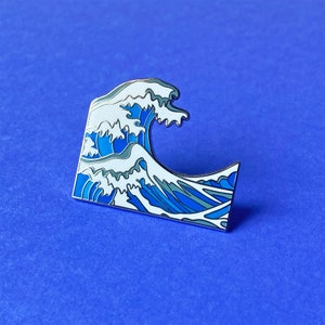 May include: A metal enamel pin shaped like a stylized ocean wave. The design features a large cresting wave with white foam and various shades of blue water, outlined in thin silver metal lines, capturing a classic Japanese art aesthetic.