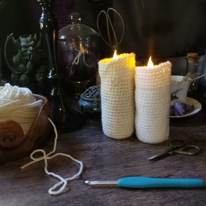 May include: Two crocheted candle covers in white, lit with a warm glow. The covers are made with a simple stitch pattern and have a rustic, handmade look. The candles are sitting on a wooden table with yarn, a crochet hook, and other craft supplies.