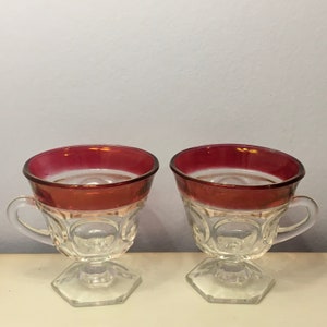 May include: Two clear glass cups featuring a wide, deep red band around the upper rim. Each cup has a circular embossed pattern on the body, a small handle on the side, and a sturdy, hexagonal base that supports the bowl of the glass vessel.