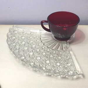 May include: A deep red glass teacup sits on a clear, fan-shaped glass snack plate. The plate features a textured, pressed starburst pattern across its surface, with a smooth, circular indentation designed to hold the base of the teacup securely.