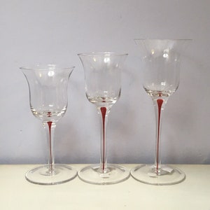 May include: Three clear glass tulip-shaped candle holders with a red stem and base. The candle holders are of varying heights, with a flared top and a round base. The design is simple and elegant.