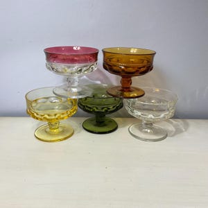 May include: A collection of vintage coupe glasses in various colors, including pink, amber, yellow, green, and clear. Each glass features a decorative, textured bowl and a stemmed base. The glasses are arranged in a stacked display.