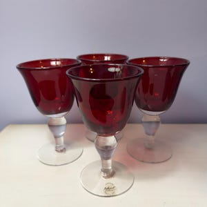 May include: A set of four wine glasses featuring deep red bowls and clear glass stems and bases. Each glass has a wide, flared rim and a rounded bowl shape. One base displays a small circular sticker with the text Ashland printed on it.