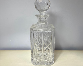 May include: A clear, faceted glass decanter with a matching stopper. The decanter has a square base and a geometric pattern. The stopper is a round, faceted shape. The overall design is elegant and classic.