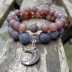 Könnte beinhalten: Zwei Armbänder mit Perlen zum Thema Himmel. Das obere Armband hat matte mauve und orangefarbene Perlen, während das untere Armband dunkelblaue und braun gemusterte Perlen hat. Ein silberner Mondsichel-Anhänger hängt am unteren Armband.