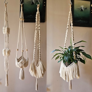 Puede incluir: Colección de colgadores de plantas de macramé hechos a mano en varios estilos. Los colgadores están hechos de cordón de algodón color crema y presentan intrincados patrones de nudos, borlas y cuentas de madera. Uno sostiene una planta en maceta con hojas verdes.