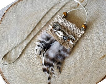 May include: A pendant necklace featuring a rectangular burlap patch adorned with a silver-toned arrow charm, a white cowrie shell, and small beads. A cluster of brown, black, and white feathers hangs from the bottom, suspended by a thin cord.