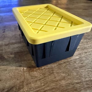 May include: A rectangular storage container with a yellow lid and a dark blue body. The lid has a grid pattern, and the container has vertical cutouts on the sides. The container is sitting on a wooden surface.