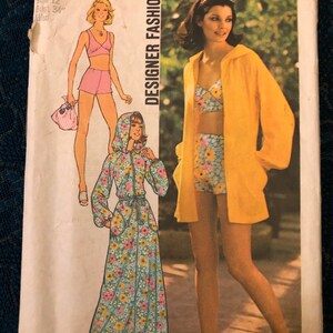 May include: A vintage Simplicity sewing pattern for a dress and a two-piece outfit. The pattern is for size 12, bust 34 inches. The dress is a long, floral print maxi dress with a hood. The two-piece outfit is a floral print top and shorts with a yellow jacket. The pattern is from the 1970s.