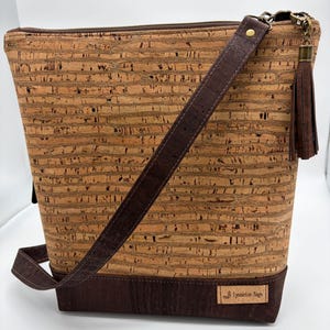 May include: A brown cork crossbody bag with a dark brown leather strap and base. The bag features a zipper closure, a decorative tassel, and a small tag that reads "LynnieLou Bags". The bag is designed with a textured, natural aesthetic.