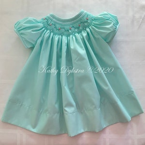 May include: A light blue smocked dress with a pink floral embroidery design around the neckline. The dress has short puffed sleeves and a gathered skirt. The dress is made of a soft, lightweight fabric.
