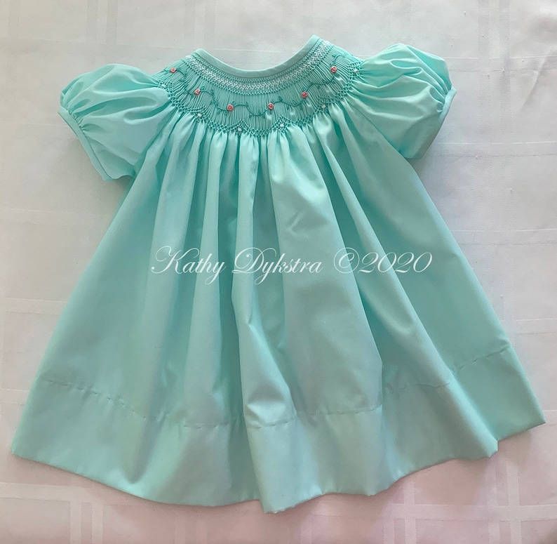May include: A light blue smocked dress with a pink floral embroidery design around the neckline. The dress has short puffed sleeves and a gathered skirt. The dress is made of a soft, lightweight fabric.