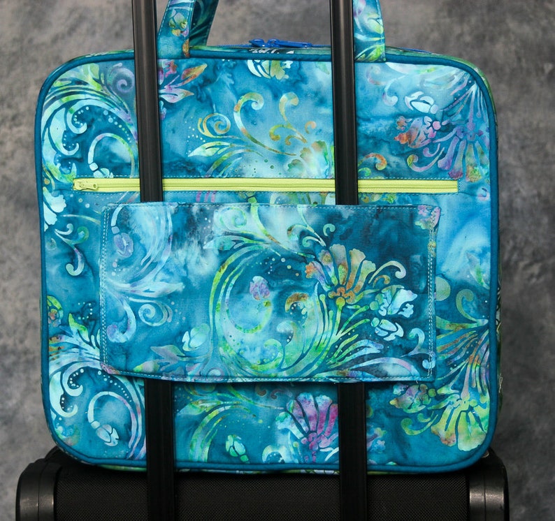 May include: A blue and green patterned travel bag with a zippered pocket and two handles. The bag is attached to a rolling luggage set.