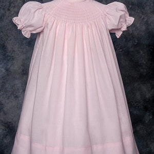May include: A pink smocked dress with short puffed sleeves and a gathered skirt. The dress has a simple, classic design and is perfect for a special occasion.