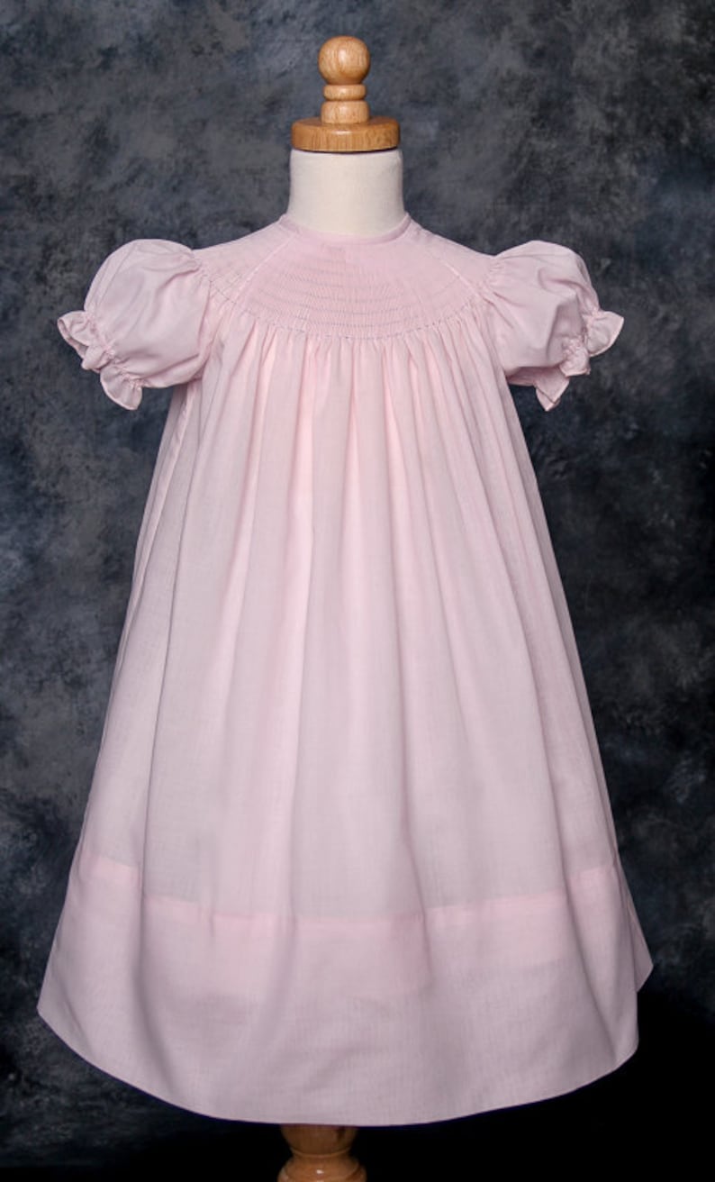May include: A pink smocked dress with short puffed sleeves and a gathered skirt. The dress has a simple, classic design and is perfect for a special occasion.