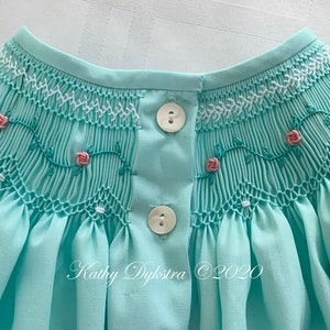 May include: A light blue smocked baby dress with white and pink embroidery of roses and vines. The dress has two white buttons on the back.
