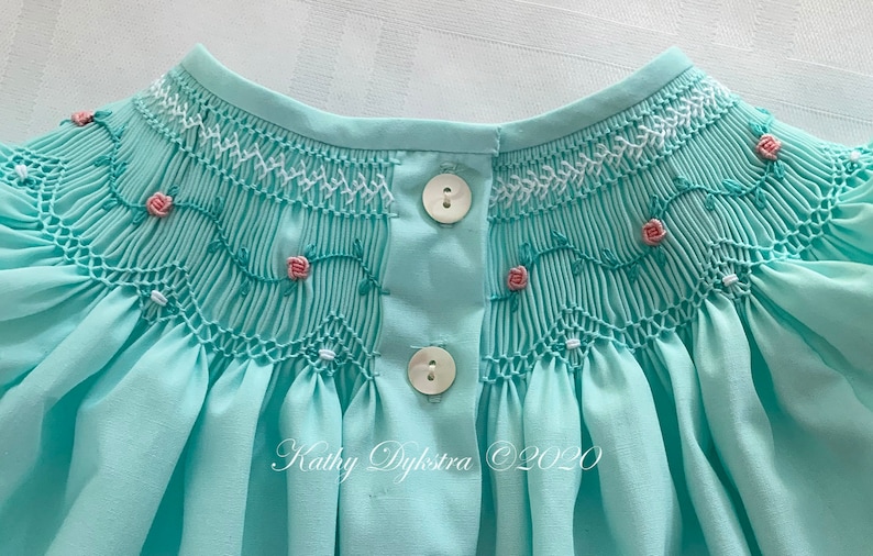 May include: A light blue smocked baby dress with white and pink embroidery of roses and vines. The dress has two white buttons on the back.
