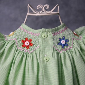May include: A green gingham baby dress with white and pink stitching. The dress has a smocked bodice with embroidered flowers and buttons. The sleeves are short and have ruffles.