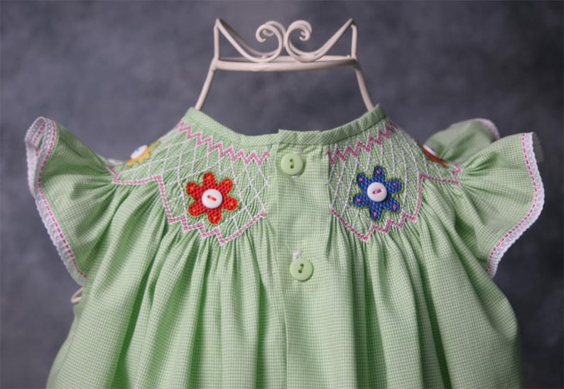 May include: A green gingham baby dress with white and pink stitching. The dress has a smocked bodice with embroidered flowers and buttons. The sleeves are short and have ruffles.