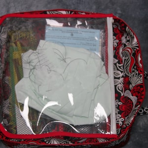 May include: A black and white patterned zippered bag with a red trim and a clear plastic window. The window shows a white piece of paper and some black string.