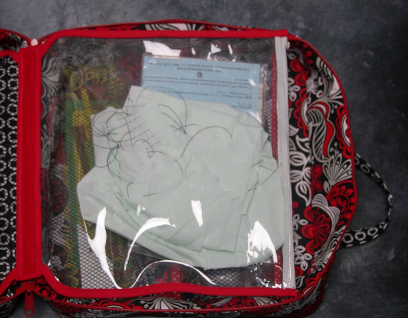 May include: A black and white patterned zippered bag with a red trim and a clear plastic window. The window shows a white piece of paper and some black string.