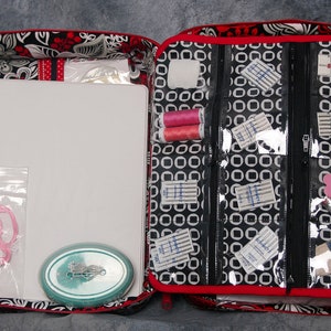 May include: Open red and black fabric tote bag with a white background and floral pattern. The bag is filled with sewing supplies, including thread, needles, and fabric scraps. The bag has a zippered compartment and a clear plastic window.