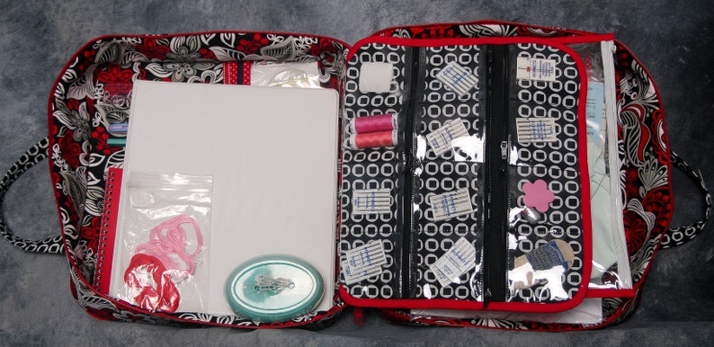 May include: Open red and black fabric tote bag with a white background and floral pattern. The bag is filled with sewing supplies, including thread, needles, and fabric scraps. The bag has a zippered compartment and a clear plastic window.