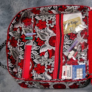 May include: A red and black floral fabric zippered pouch with multiple pockets and compartments. The pouch is open and filled with various items, including pens, a notebook, a roll of tape, and adhesive bandages.