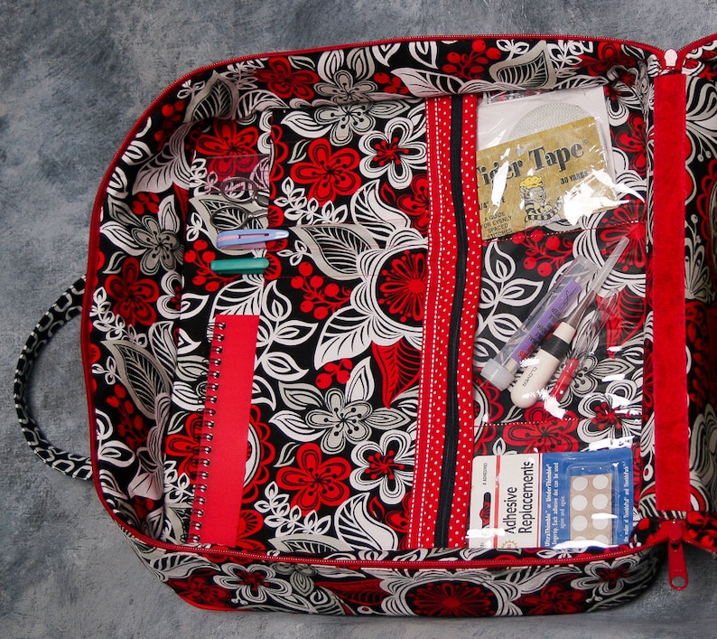 May include: A red and black floral fabric zippered pouch with multiple pockets and compartments. The pouch is open and filled with various items, including pens, a notebook, a roll of tape, and adhesive bandages.