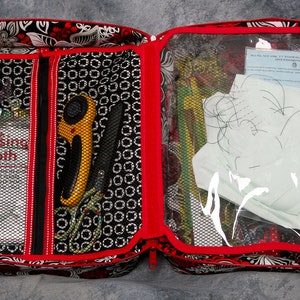 May include: Open red and black fabric sewing bag with a clear zippered pouch. The pouch contains white fabric, a piece of paper, and a sewing needle. The bag has a mesh pocket with a label that says "Rotary Pressing Cloth".