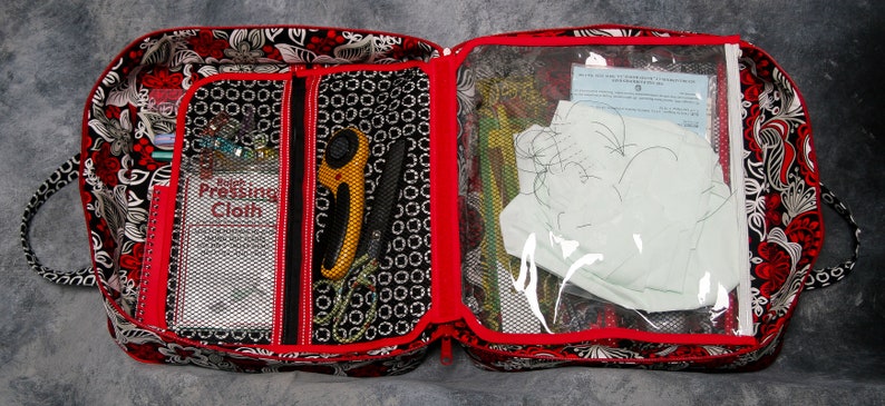 May include: Open red and black fabric sewing bag with a clear zippered pouch. The pouch contains white fabric, a piece of paper, and a sewing needle. The bag has a mesh pocket with a label that says "Rotary Pressing Cloth".