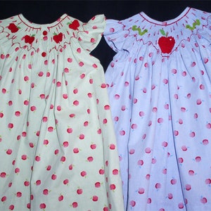 May include: Two girls' dresses with apple print and embroidered apple details. One dress is light green with red apples and the other is light blue with red apples.