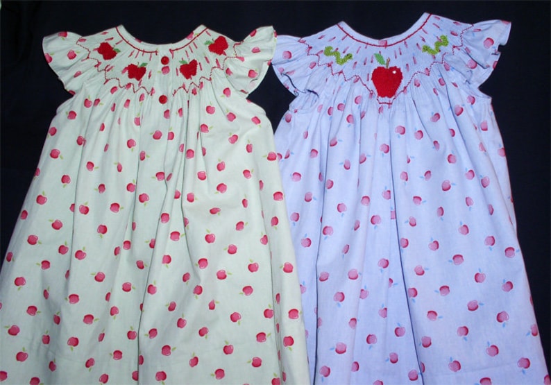 May include: Two girls' dresses with apple print and embroidered apple details. One dress is light green with red apples and the other is light blue with red apples.