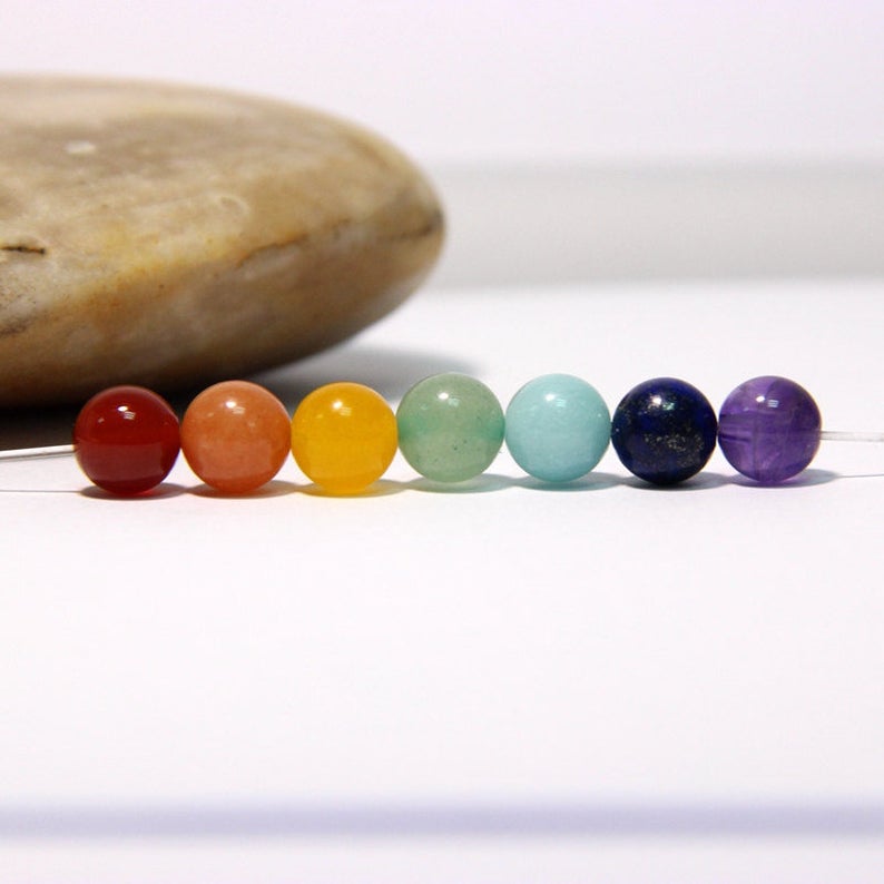 May include: A row of seven round gemstone beads in the colors of the rainbow, from red to purple. The beads are arranged in a line on a white surface.
