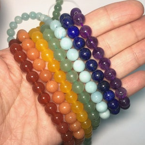May include: Seven chakra bracelets made of natural stones in a rainbow color pattern. The bracelets are stacked on top of each other, starting with red, orange, yellow, green, blue, indigo, and violet.