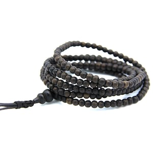 Puede incluir: Un collar de cuentas de oración de madera marrón oscuro, también conocido como mala, con una borla. Las cuentas son pequeñas y redondas, ensartadas para formar múltiples hebras. El collar se muestra sobre un fondo blanco. Las cuentas miden unos 6 mm de diámetro.
