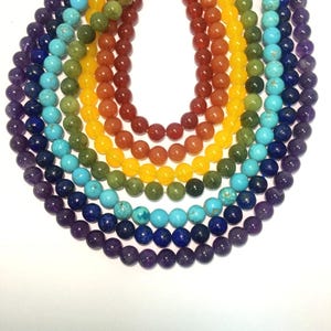 May include: A set of seven chakra gemstone beads in a rainbow color pattern. The beads are arranged in a circle, with the purple amethyst beads at the bottom, followed by blue lapis lazuli, turquoise, green aventurine, yellow citrine, orange carnelian, and red jasper beads.