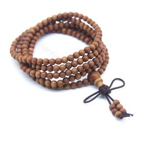 Puede incluir: Una pulsera de cuentas de oración de madera con múltiples hilos de pequeñas cuentas redondas marrones. La pulsera tiene una cuenta decorativa de madera y una borla anudada marrón oscuro. Las cuentas están ensartadas, creando un efecto en capas.