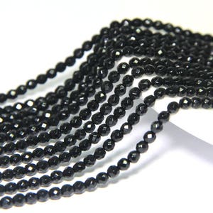 Puede incluir: Primer plano de múltiples hebras de pequeñas cuentas negras facetadas. Las cuentas están ensartadas, creando una superficie texturizada y reflectante. Las cuentas son de un negro profundo y brillante, y las facetas captan la luz, creando un efecto brillante.