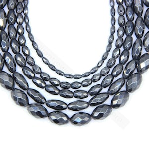 May include: Close-up of multiple strands of faceted, dark grey hematite gemstone beads. The beads are oval-shaped with a polished, reflective surface. The strands vary in size, creating a layered effect. The background is white.