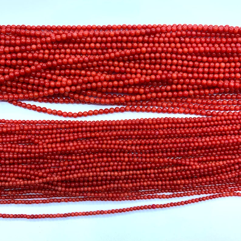 May include: Close-up of multiple strands of red coral beads. The beads vary in size, with the largest at the top and the smallest at the bottom. The beads are a vibrant, uniform red color, and are strung together, creating a textured surface.