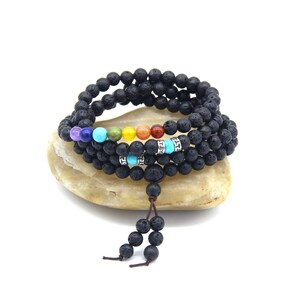 Op de afbeelding: Zwarte lavasteen kralenarmbanden met kleurrijke chakra kralen. De armbanden zijn gestapeld op een lichtgekleurde steen. De chakra kralen zijn in de kleuren van de regenboog, met een kwast van zwarte kralen.